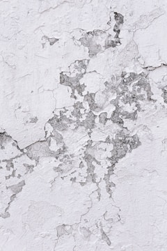 Cracked and peeling white paint on a textured surface reveals patches of dull gray material beneath. The uneven layers create a rugged and weathered appearance.