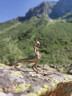 A small metallic figurine resembling a creature with a long tail stands on a rocky surface. The background features a lush, green mountain under a clear blue sky, suggesting a serene natural setting.