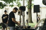 A DJ is performing outdoors, wearing headphones and operating a DJ controller with speakers and audio equipment nearby. The scene is set in a park with trees and a grass area visible in the background. Another person stands nearby, wearing a casual outfit and a hat. A few people can be seen in the distance, and the atmosphere appears relaxed.