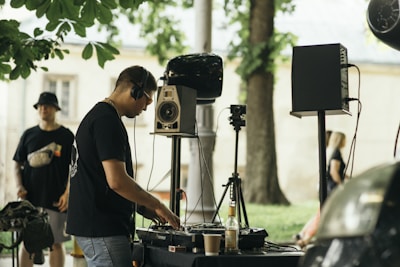 A DJ is performing outdoors, wearing headphones and operating a DJ controller with speakers and audio equipment nearby. The scene is set in a park with trees and a grass area visible in the background. Another person stands nearby, wearing a casual outfit and a hat. A few people can be seen in the distance, and the atmosphere appears relaxed.