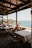 A serene outdoor dining area with several wooden tables and chairs, all covered with white tablecloths. The area is shaded by a wooden pergola, and overlooks a beautiful coastal view with clear blue water, distant greenery, and a rocky shore. Flower pots with red flowers are placed along the edge of the dining area.
