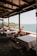 A serene outdoor dining area with several wooden tables and chairs, all covered with white tablecloths. The area is shaded by a wooden pergola, and overlooks a beautiful coastal view with clear blue water, distant greenery, and a rocky shore. Flower pots with red flowers are placed along the edge of the dining area.