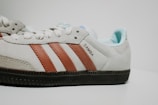 A close-up view of a suede sneaker with a light beige color and brown leather stripes. The word 'SAMBA' is printed on the side, and the shoe features a classic design with a rubber sole.