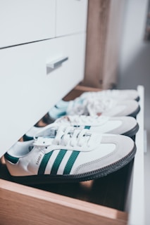 A pair of white sneakers with green stripes are neatly placed inside an open wooden drawer. The sneakers display a clean and stylish design, and the word 'SAMBA' is visible on the side. The background features a white cabinet and a blurred interior setting.