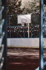 A basketball court with a hoop and backboard is surrounded by a high fence decorated with colorful triangular flags. The court surface is a deep maroon, framed by tall green trees in the background creating a serene and somewhat secluded atmosphere.