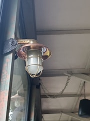 An outdoor light fixture with a metallic cage surrounding a frosted glass bulb is mounted on a wall. The structure appears rustic with visible signs of weathering. Above, the ceiling is supported by metal beams and has chains hanging, contributing to an industrial aesthetic.