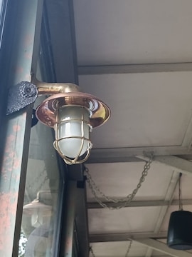 An outdoor light fixture with a metallic cage surrounding a frosted glass bulb is mounted on a wall. The structure appears rustic with visible signs of weathering. Above, the ceiling is supported by metal beams and has chains hanging, contributing to an industrial aesthetic.