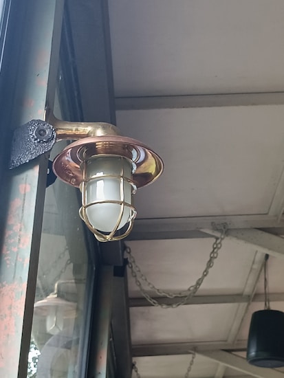 An outdoor light fixture with a metallic cage surrounding a frosted glass bulb is mounted on a wall. The structure appears rustic with visible signs of weathering. Above, the ceiling is supported by metal beams and has chains hanging, contributing to an industrial aesthetic.