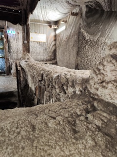 The interior features walls and ceilings covered with salt or a mineral-like substance, creating a textured, crystalline appearance. There is a wooden structure in the middle that could be a walkway or railing. Soft lighting provides a subtle glow, highlighting the rugged surface details.