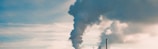 Large industrial chimneys emit thick clouds of smoke into the sky. The smoke is dark and contrasts with the light, partially cloudy sky in the background.