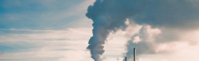 Large industrial chimneys emit thick clouds of smoke into the sky. The smoke is dark and contrasts with the light, partially cloudy sky in the background.