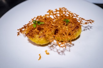 A dish featuring two golden croquettes set on a white plate, topped with an intricate lattice of crispy cheese and garnished with small sprigs of fresh green herbs.