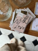 A stack of greeting cards with a prominent card displaying the words 'With Love' in bold, black script. The cards are neatly arranged on a wooden surface, next to a large, transparent glass vessel. In the background, part of a patterned floor is visible, with a person standing that appears slightly blurred.
