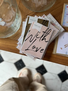 A stack of greeting cards with a prominent card displaying the words 'With Love' in bold, black script. The cards are neatly arranged on a wooden surface, next to a large, transparent glass vessel. In the background, part of a patterned floor is visible, with a person standing that appears slightly blurred.