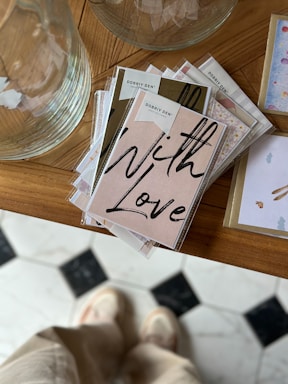 A stack of greeting cards with a prominent card displaying the words 'With Love' in bold, black script. The cards are neatly arranged on a wooden surface, next to a large, transparent glass vessel. In the background, part of a patterned floor is visible, with a person standing that appears slightly blurred.