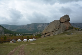 A large rock formation dominates the landscape, with several white circular yurts nearby on a grassy plain. The area is surrounded by mountains covered with greenery and patches of autumn foliage. The sky is overcast, creating a moody ambiance.