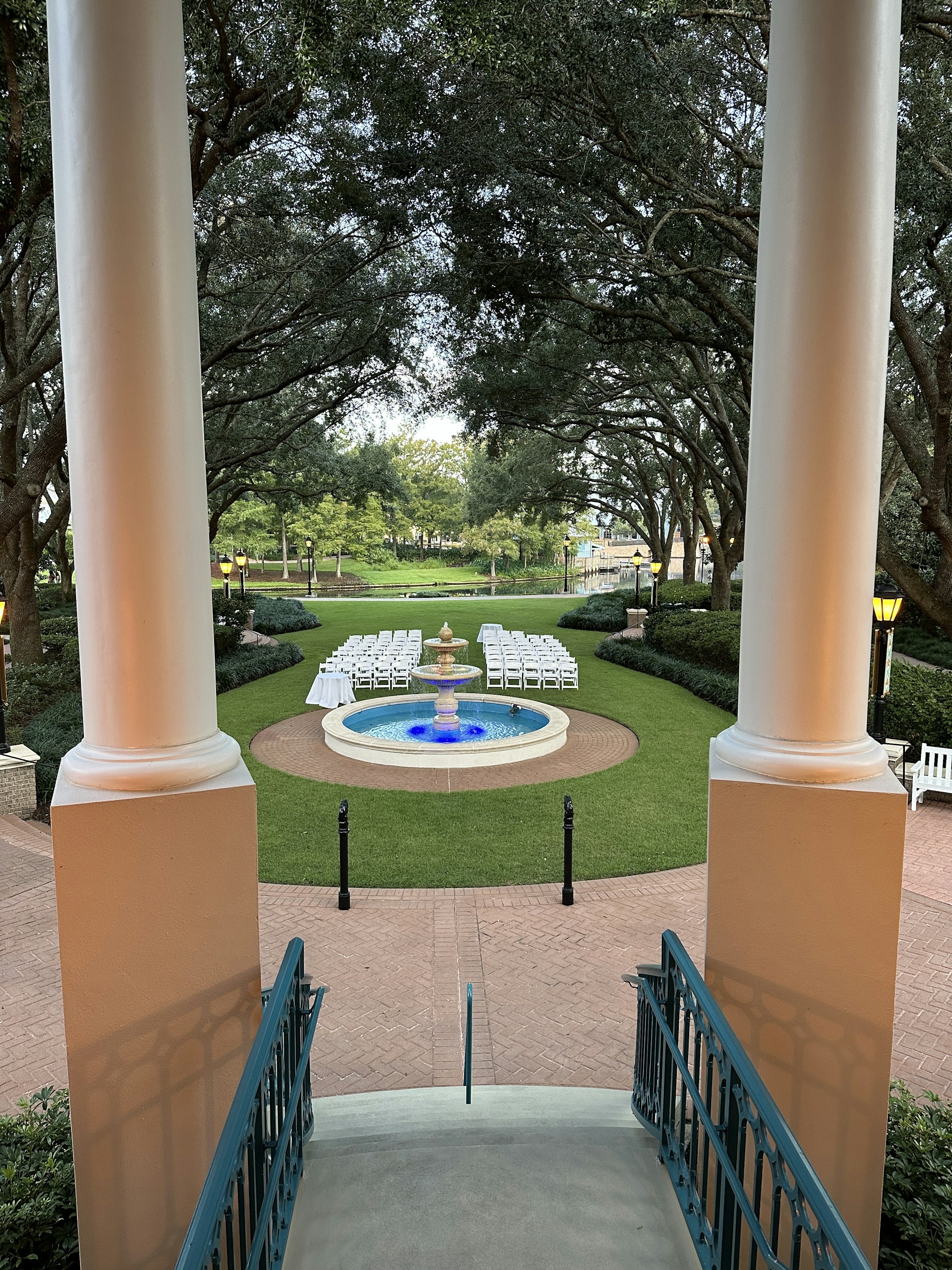 View from a staircase leading down to a garden area with symmetrical rows of white chairs facing a central fountain surrounded by green grass and trees. The setting features tall columns and lantern-style lights, creating an elegant atmosphere.