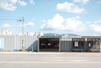 A modern bus stop is situated on a quiet street with a sign reading 'Chuncheon Station'. In the background, residential high-rise buildings and a mountainous landscape are visible. The foreground features an electric scooter parked near the stop. The sky is bright and mostly clear, filled with scattered fluffy clouds.
