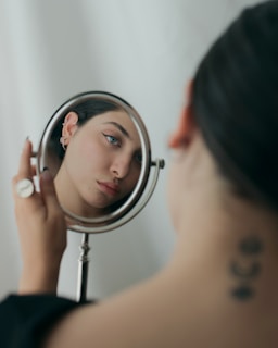 A person gazes at their reflection in a handheld circular mirror. The focus is on the face, with minimal expression and a serene appearance. The background is blurred, emphasizing the face in the mirror. The individual has dark hair and is wearing a black top. A tattoo with three crescent shapes is visible on the back of the neck.