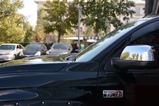 A shiny black vehicle is prominently displayed in the foreground with a visible badge indicating a 5.7L V8 engine. The side mirror reflects trees and a partially cloudy sky, suggesting an outdoor setting. In the background, there are other vehicles parked side by side, along with greenery and a building, likely in a parking lot.
