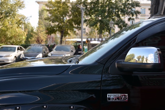 A shiny black vehicle is prominently displayed in the foreground with a visible badge indicating a 5.7L V8 engine. The side mirror reflects trees and a partially cloudy sky, suggesting an outdoor setting. In the background, there are other vehicles parked side by side, along with greenery and a building, likely in a parking lot.