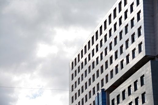 A large modern building with a grid of windows is viewed from a side angle, reflecting light gray clouds in the sky.