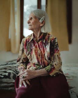 An elderly woman with grey hair sits on a bed, looking out a window to her left. She wears a colorful, patterned blouse and a maroon skirt. A pearl hair clip adorns her hair. The room is softly lit with neutral-colored curtains and a patterned blanket visible in the background.