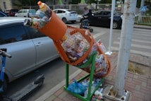 A tilted, overfilled recycle bin designed in the shape of a rocket, mainly filled with plastic bottles and other recyclables, is situated near a sidewalk. It is supported by a green metal stand on a paved surface, with some additional waste scattered around it. In the background, a street with parked cars and a person riding a scooter can be seen.