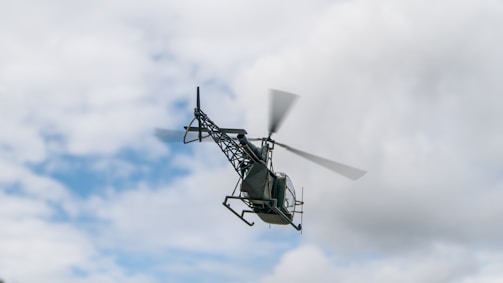 A helicopter is seen flying against a backdrop of cloudy sky. The helicopter appears to be mid-flight, with its rotor blades spinning rapidly, suggesting motion through the air.