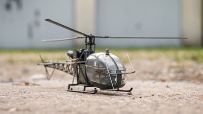 A small model helicopter rests on a textured ground, with its metallic and plastic components visible. The backdrop is slightly blurred, suggesting an outdoor setting. The helicopter model appears detailed, with a prominent rotor blade and intricate framework beneath the main body.