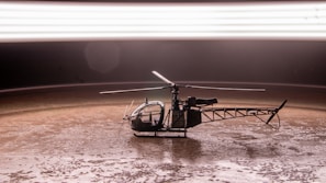 A small helicopter model placed on a textured brown surface under a bright, blurred horizontal light streak. The helicopter is detailed with visible rotors and a clear cockpit.