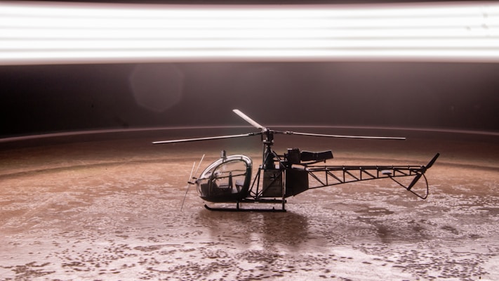 A small helicopter model placed on a textured brown surface under a bright, blurred horizontal light streak. The helicopter is detailed with visible rotors and a clear cockpit.