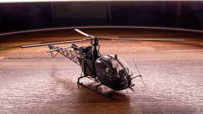 A detailed model helicopter with a visible cockpit and intricate rotor blades is placed on a wooden surface. The lighting highlights the textures of both the helicopter and the wood, creating a dynamic contrast between the metal and wooden elements.