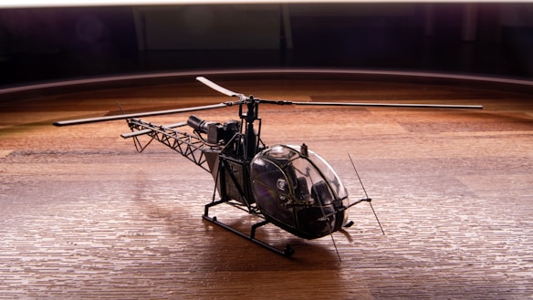 A detailed model helicopter with a visible cockpit and intricate rotor blades is placed on a wooden surface. The lighting highlights the textures of both the helicopter and the wood, creating a dynamic contrast between the metal and wooden elements.