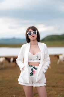 A person in a stylish white outfit with floral patterns stands confidently outdoors. They sport large, fashionable sunglasses and have short brown hair. In the background, there are cows grazing in an open field with a water body and hills visible at a distance under a cloudy sky.
