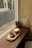 A cozy café scene with a wooden table by a window, featuring a cup of latte art coffee and a piece of cake with chocolate shavings served on a wooden tray. A small potted plant adds a touch of greenery. The exterior view shows a street with scattered leaves and a few shops with glass fronts.