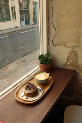 A cozy caf&eacute; scene with a wooden table by a window, featuring a cup of latte art coffee and a piece of cake with chocolate shavings served on a wooden tray. A small potted plant adds a touch of greenery. The exterior view shows a street with scattered leaves and a few shops with glass fronts.