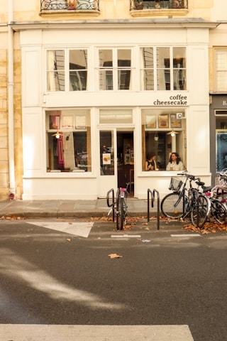 A cozy café storefront displays a sign that reads 'coffee cheesecake'. Two people can be seen sitting inside through large windows, with bicycles parked outside. The building features a cream facade with decorative window grilles above.