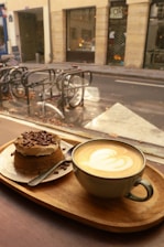 A warm, cozy cafe setting with a latte featuring a heart-shaped design in the foam, served on a wooden tray alongside a small chocolate cake topped with cream and chocolate shavings. Outside the window, there are bicycles parked on a street with a muted, autumnal atmosphere.