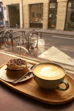 A warm, cozy cafe setting with a latte featuring a heart-shaped design in the foam, served on a wooden tray alongside a small chocolate cake topped with cream and chocolate shavings. Outside the window, there are bicycles parked on a street with a muted, autumnal atmosphere.