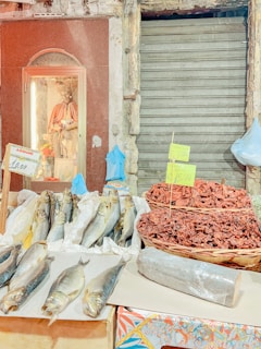 A market stall displays a variety of goods, including fresh fish laid out on paper and dried goods in a woven basket. There is a statue of a saint or religious figure in a small alcove with a glass front. The background shows a metal shutter and old stonework. Price signs are visible on some items.