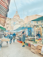 A vibrant street market with various stalls selling fresh produce and clothes. People are walking and shopping under colorful awnings. A historic building with intricate architecture is visible in the background.
