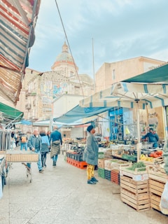 A vibrant street market with various stalls selling fresh produce and clothes. People are walking and shopping under colorful awnings. A historic building with intricate architecture is visible in the background.