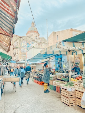 A vibrant street market with various stalls selling fresh produce and clothes. People are walking and shopping under colorful awnings. A historic building with intricate architecture is visible in the background.