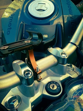 A close-up view of a motorcycle's handlebars and upper structures. Visible components include the handle grips, a metallic clamp with a black mount, the ignition key slot, and part of the dashboard. The fuel tank cap with a lock is prominently in the frame, with 'Regulmoto' branding visible. The color tones are metallic and convey a robust, industrial feel.
