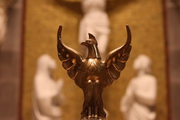 A bronze or gold-colored sculpture of a bird with wings spread, likely symbolizing a phoenix, positioned in the foreground with blurred white statues or figures in the background. The backdrop appears to be a textured, possibly gilded surface.