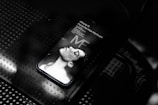 A smartphone displaying a grayscale image of a person lying down with eyes closed. The words 'iPhone 15 Pro or Pro Max Mockups' are visible on the screen. The phone rests on a perforated metallic surface.