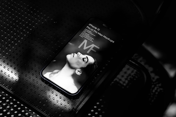 A smartphone displaying a grayscale image of a person lying down with eyes closed. The words 'iPhone 15 Pro or Pro Max Mockups' are visible on the screen. The phone rests on a perforated metallic surface.