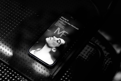 A smartphone displaying a grayscale image of a person lying down with eyes closed. The words 'iPhone 15 Pro or Pro Max Mockups' are visible on the screen. The phone rests on a perforated metallic surface.