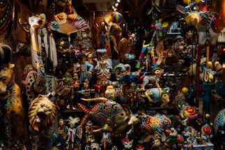 A crowded display of colorful, intricately painted ceramic and wooden figurines, including animals, mythical creatures, and human figures. The items exhibit a mix of vibrant patterns and designs, creating a lively and festive atmosphere.